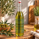 A clear glass swing-top bottle filled with olive oil stands on a wooden table, surrounded by olives, olive branches, a small secondary bottle, and kitchen decor in a warm indoor setting.
