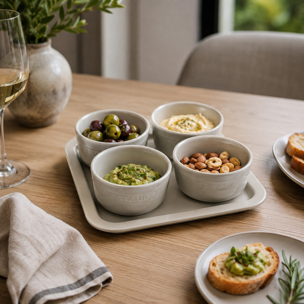 Four small round ceramic bowls arranged on a rectangular tray on a wooden table, filled with olives, hummus, guacamole, and nuts. A wine glass, napkin, and bread are visible in a styled dining setting.