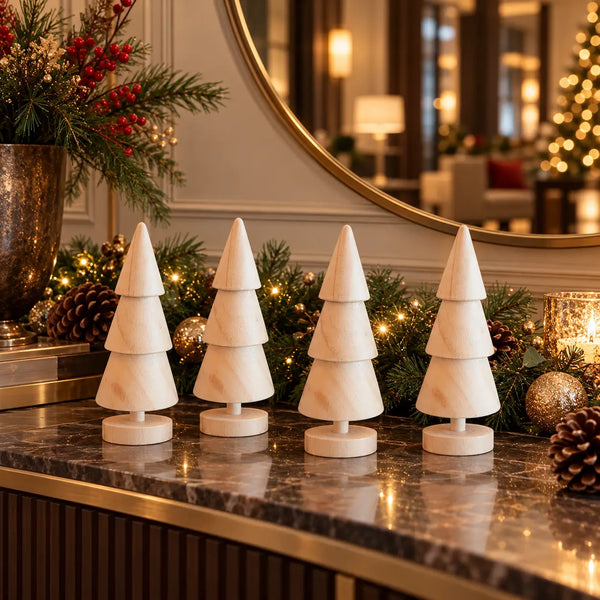 Four small white wooden Christmas tree ornaments with tiered designs are arranged on a dark marble console table in a decorated holiday setting with pinecones, garland, and warm lights.