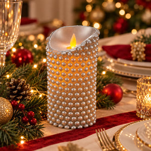A decorative pillar candle wrapped in rows of white faux pearls glows with a warm yellow flame, set on a holiday table runner surrounded by pine branches, red ornaments, and gold tableware.