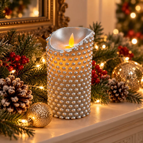A silver bead-covered pillar candle with a glowing flame sits on a mantel surrounded by Christmas greenery, pinecones, red berries, and warm string lights.