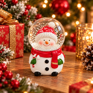 A Christmas snow globe figurine shaped like a smiling snowman, wearing a red Santa hat, red scarf, and green mittens, displayed on a wooden table with festive holiday decorations in the background.