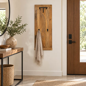 A rustic wooden entryway coat rack with black metal hooks hangs on a cream wall in a styled home entryway, with a beige throw blanket draped over it.