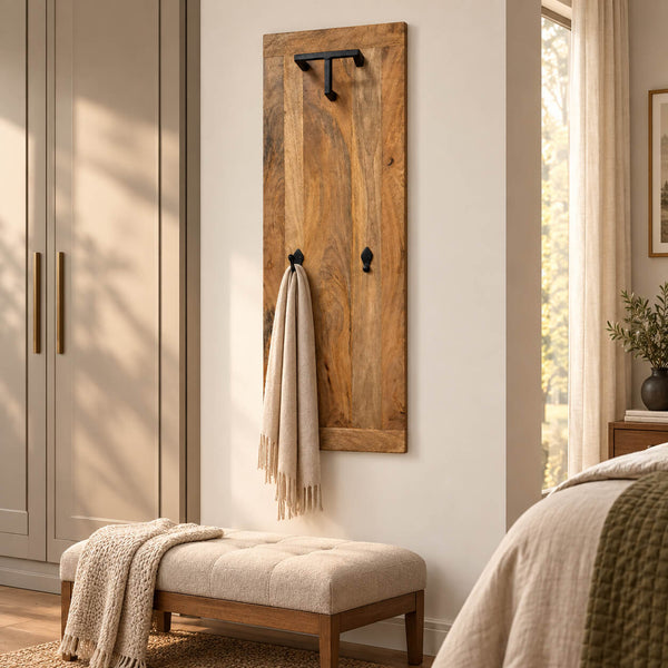 A rustic wooden wall-mounted coat rack with black metal hooks, styled in a warm, modern bedroom setting next to a tufted bench and window.