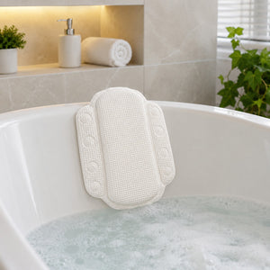 A white textured bath cushion pad with snap fasteners, shown hanging on the rim of a white bathtub filled with bubbly water in a bright, modern bathroom setting.