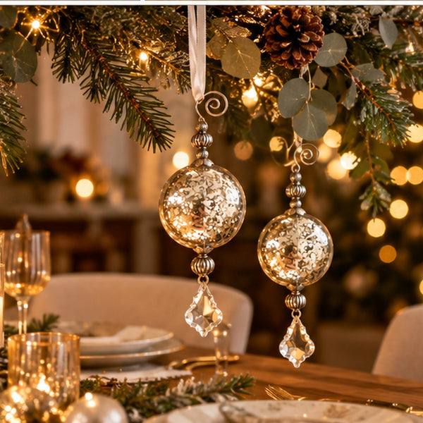Two large hammered silver glass Christmas tree ornaments hang from a decorated holiday garland with pine branches, eucalyptus leaves, and warm string lights above a dining table.