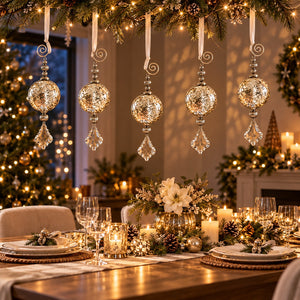 A beautifully decorated Christmas dining table with hanging silver crystal ornaments, warm string lights, candles, floral centerpieces, and place settings, with a decorated Christmas tree and fireplace in the background.