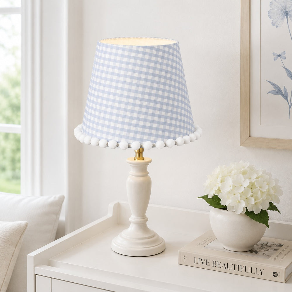 Micasa pink gingham floral lampshade with pom-pom trim displayed on a bedside table in a pastel children’s bedroom.