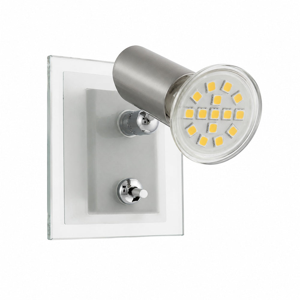 A modern wall-mounted spotlight lamp with a brushed silver metal head and clear acrylic backplate, shown lit in a bedroom setting next to a bed with a gray headboard.