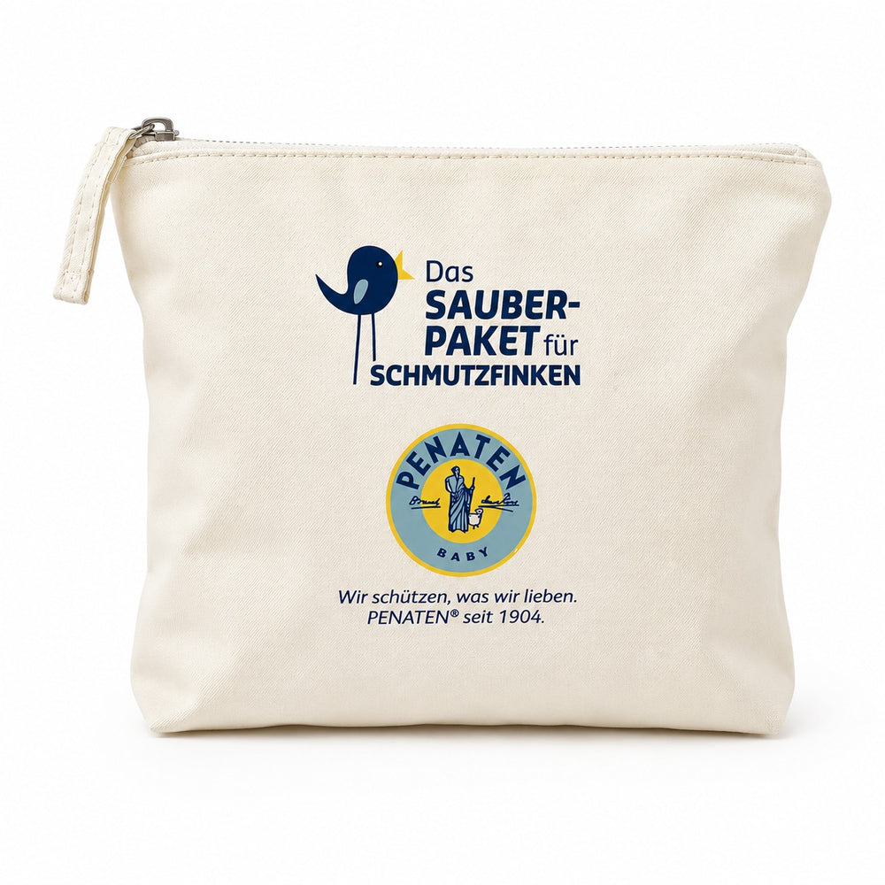 A cream-colored canvas pouch with printed branding for 'Das Sauber-Paket für Schmutzfinken' and 'PENATEN BABY' sits on a light wooden table surrounded by baby-themed props like a plush bunny, folded towels, and a bottle, creating a warm, soft-focus lifestyle setting.