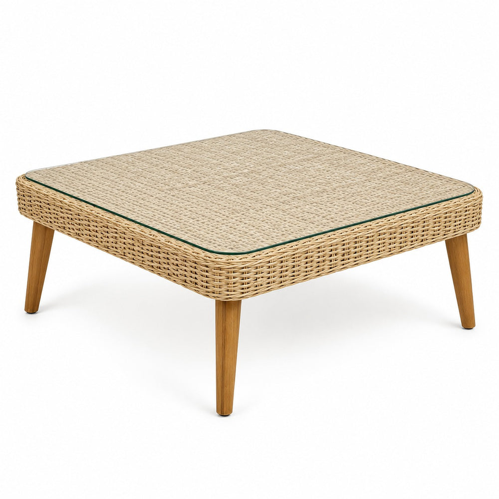 A square woven rattan coffee table with a clear glass top and light wood legs, styled in a cozy living room next to a beige sofa and an olive green armchair.