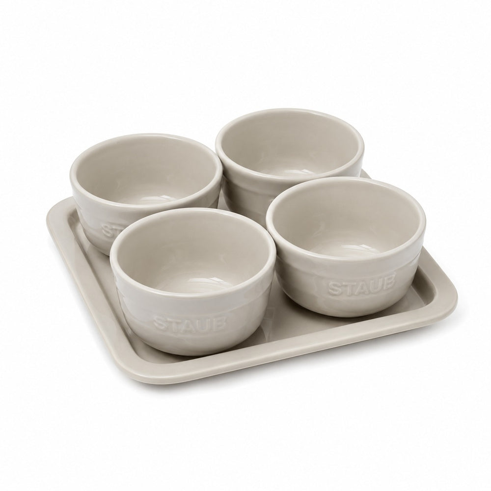 Four small round ceramic bowls arranged on a rectangular tray on a wooden table, filled with olives, hummus, guacamole, and nuts. A wine glass, napkin, and bread are visible in a styled dining setting.