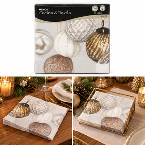 Migros Cucina e Tavola paper napkins with a printed design of metallic gold and white Christmas ornaments, shown in its retail packaging and styled on a wooden table with candles and tableware.