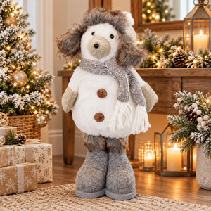 A plush dog-shaped Christmas decoration wearing a white coat, gray furry hat and scarf, and gray boots, standing in a warmly lit holiday room with a decorated tree, candles, and wrapped gifts.