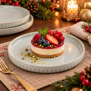 A small cheesecake dessert topped with fresh berries, blueberries, a strawberry slice, and a mint leaf, served on a light blue plate in a cozy holiday table setting with candles, pinecones, and a gold fork.