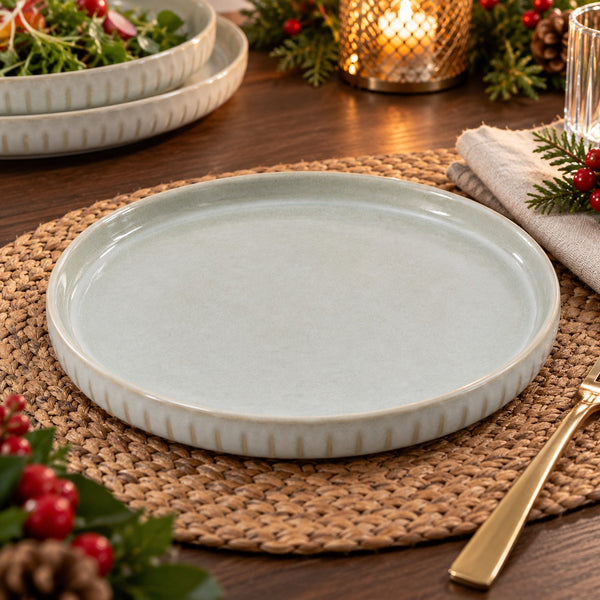 A round ceramic plate with a pale sage green center and a warm beige flared rim, styled on a woven placemat on a wooden table with holiday greenery and a candle in the background.