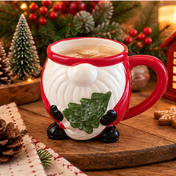 A festive ceramic mug shaped like a gnome with a red rim and handle, a white face and body, black shoes, and a green holly tree on the chest. The mug is filled with a hot beverage and set on a wooden surface with Christmas decorations in the background.