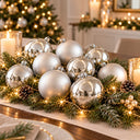 A festive Christmas table centerpiece featuring a cluster of shiny silver and matte white baubles nestled in green pine garland with warm white string lights, pinecones, and candles in a softly blurred holiday room setting.