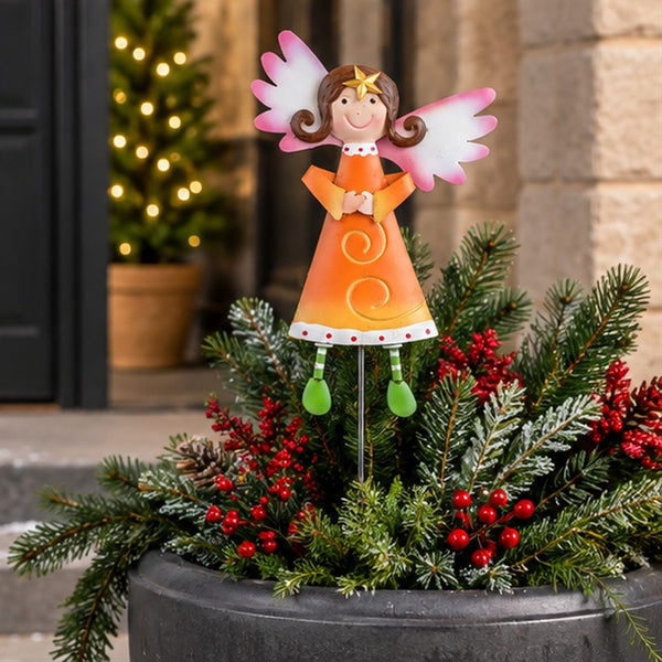 A decorative angel garden stake with a smiling angel figure in an orange dress and pink wings, placed in a planter filled with green pine branches and red berries in front of a doorway.