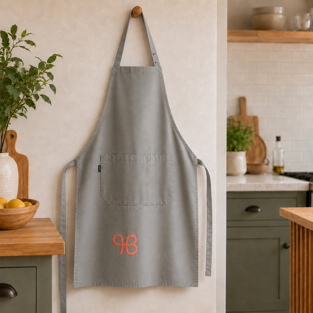 A light gray kitchen apron with a single front pocket and orange embroidered initials, hanging on a wooden hook in a kitchen setting.