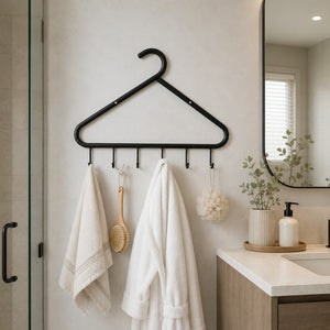A black metal hanger-style robe rack with five hooks hangs on a light beige bathroom wall. Two white towels, a wooden scrubber, and a white bath bomb are hanging from the hooks.