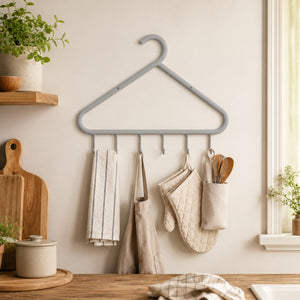 A gray metal kitchen towel hanger mounted on a beige wall, holding several kitchen towels, an apron, oven mitts, and utensils in a softly styled kitchen setting.