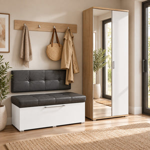 A modern entryway scene showing a white storage bench with a black tufted seat cushion, styled with a wooden coat rack, hanging bag, and beige jacket. A tall white and wood wardrobe stands beside a window, and a woven rug covers the floor.