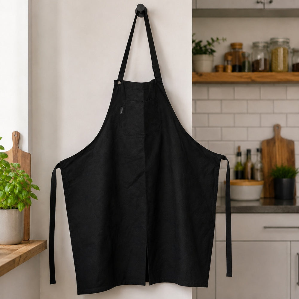 A black kitchen apron hangs from a wall hook in a bright kitchen setting, with white subway tiles, wooden shelves, and kitchen items visible in the background.