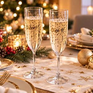 Two clear champagne flutes filled with bubbly champagne, printed with the word 'Cheers!' in black script, set on a decorated holiday table with gold star placemats and warm bokeh lights in the background.