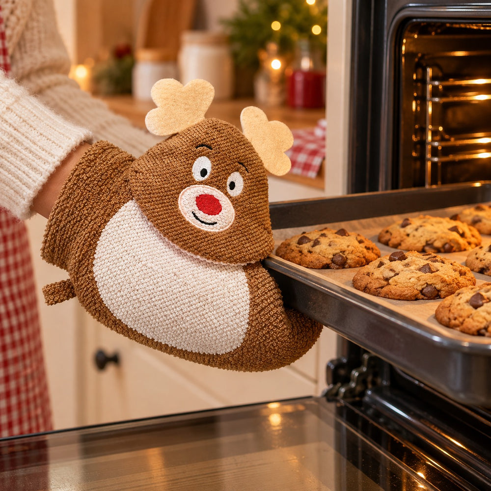 A person wearing a cream knit sweater uses a brown and white knitted reindeer-shaped oven mitt to pull a baking sheet of chocolate chip cookies out of a lit oven in a cozy kitchen setting.