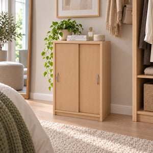 A light natural wood two-door storage cabinet with silver handles, styled in a bedroom or closet area with a potted plant on top, books, and surrounding home decor.