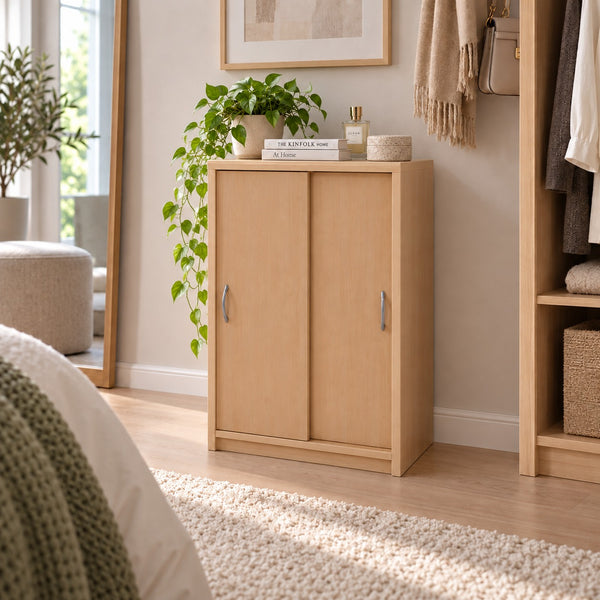 A light natural wood two-door storage cabinet with silver handles, styled in a bedroom or closet area with a potted plant on top, books, and surrounding home decor.