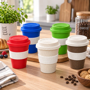 Five reusable coffee cups with screw-on lids and beige bands are arranged on a wooden table in a bright kitchen setting. The cups come in red, blue, green, and dark brown colors, with one cup showing a light beige interior.