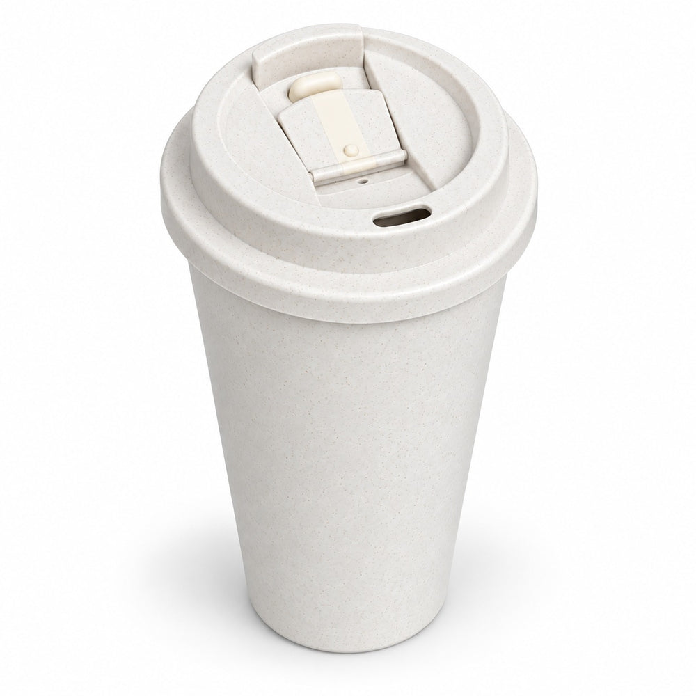 A light beige reusable travel mug with a flip-top lid sits on a wooden desk surrounded by a laptop, notebook, pen, book, and potted plant.