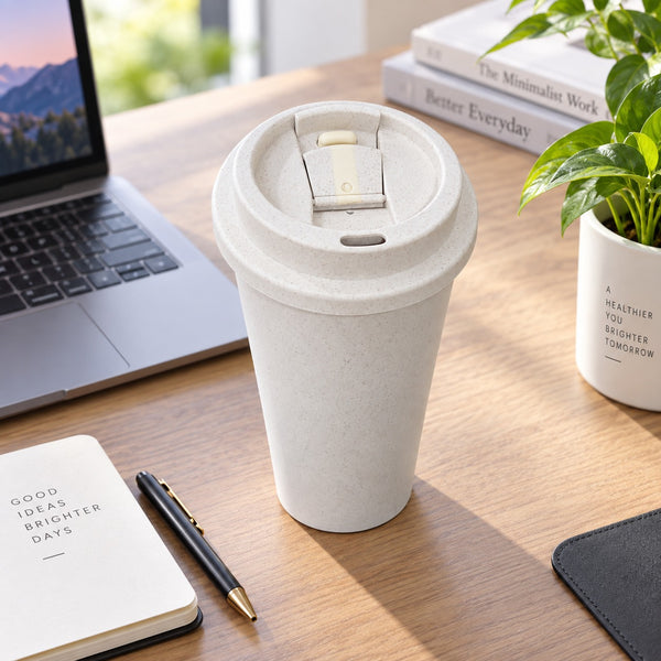 A light beige reusable travel mug with a flip-top lid sits on a wooden desk surrounded by a laptop, notebook, pen, book, and potted plant.