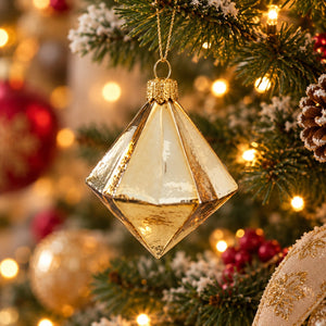A faceted, diamond-shaped gold Christmas ornament hanging from a decorated Christmas tree with warm string lights and pinecones.