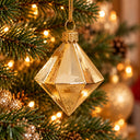 A faceted gold Christmas ornament hanging from a green Christmas tree branch with warm string lights in the background.