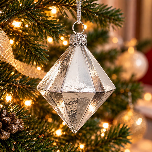 A faceted silver Christmas ornament hanging from a decorated evergreen tree with warm string lights.