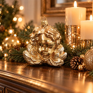 A shiny gold Christmas ornament shaped like a cherub's face sits on a wooden mantel decorated with green pine branches, pinecones, lit candles, and a blurred Christmas tree in the background.