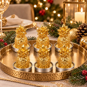 Six decorative golden pineapple candle holders arranged on a hammered round gold tray, styled with pine branches, a lit candle, and holiday lights in the background.