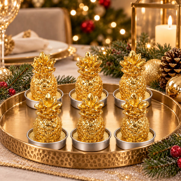 Six decorative golden pineapple candle holders arranged on a hammered round gold tray, styled with pine branches, a lit candle, and holiday lights in the background.