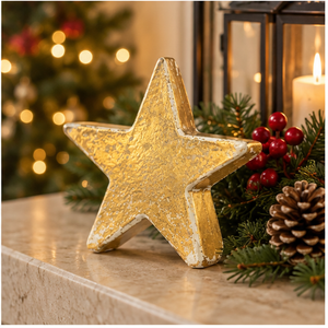 A decorative gold star ornament with a distressed, textured finish stands on a marble mantel next to holiday greenery, red berries, a pinecone, and a lit candle, with a blurred Christmas tree in the background.