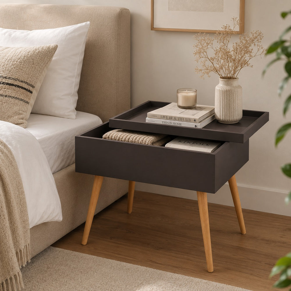 A dark charcoal gray bedside table with light wood legs, styled next to a bed with white linens. The table features a top tray holding books, a candle, and a vase, and an open drawer containing a folded knit blanket.