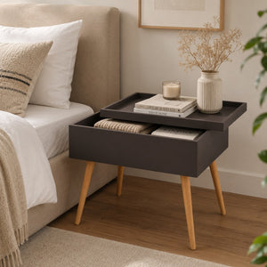 A dark charcoal gray bedside table with light wood legs, styled next to a bed with white linens. The table features a top tray holding books, a candle, and a vase, and an open drawer containing a folded knit blanket.