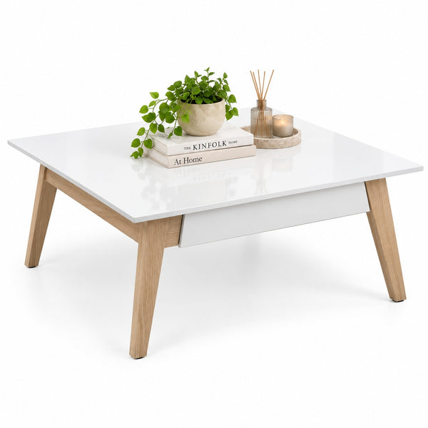 A modern low coffee table with a glossy white rectangular top, a central drawer, and light natural wood legs, styled with a potted plant, books, and a reed diffuser on a white background.
