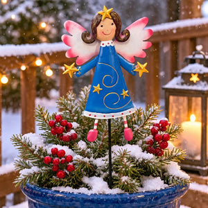 A decorative Christmas angel garden stake with a blue dress, pink wings, and gold stars, displayed in a snowy blue pot with evergreen branches and red berries on a wooden deck at night.
