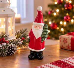 A small Santa Claus figurine stands on a wooden table in a decorated holiday setting, with a glowing Christmas tree, lantern, pine branches, and a wrapped gift box in the background.