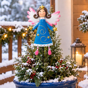 A decorative Christmas angel figurine with a blue dress, pink wings, and gold stars, displayed on a snowy evergreen tree in a blue planter. The scene is set outdoors on a porch with warm lights and candles in the background.