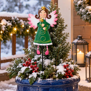 A festive holiday garden decoration featuring a painted angel figurine with pink wings and boots, standing in a snowy blue planter with greenery and red berries.
