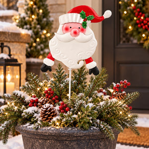 A decorative Santa Claus yard stake with a smiling white beard, red hat, and green holly accent, displayed in a snowy evergreen planter with pinecones and red berries in front of a holiday door.